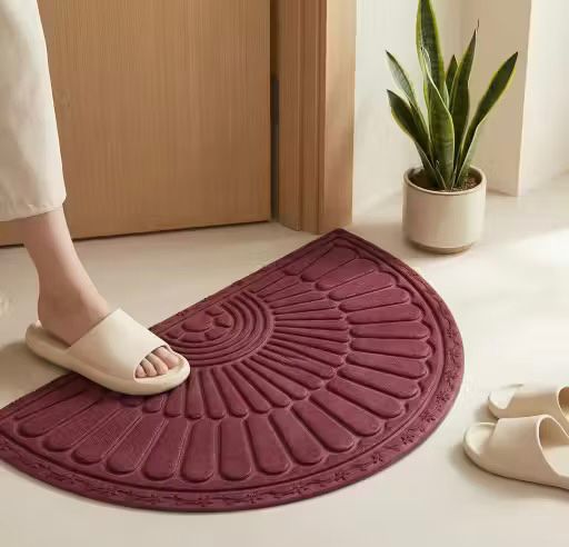 Person wearing a sandal stepping onto a burgundy bath mat with a plant and slippers in the background.
